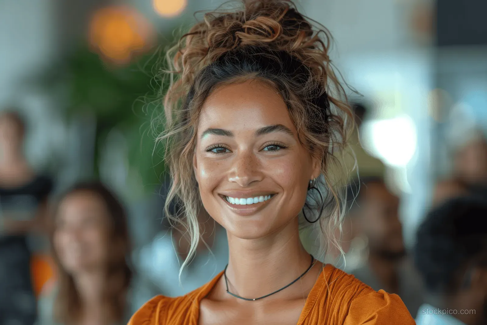 A smiling young woman with a curly hair bun and orange top looks at the camera indoors in warm light.