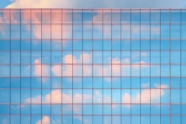 Glass facade reflects blue sky and pink-and-white clouds in a grid pattern during sunset.