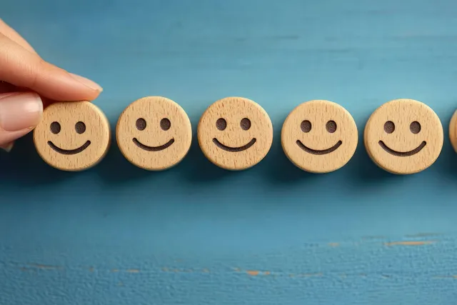A hand holds a happy wooden face token among other smiley wooden icons on a blue background.