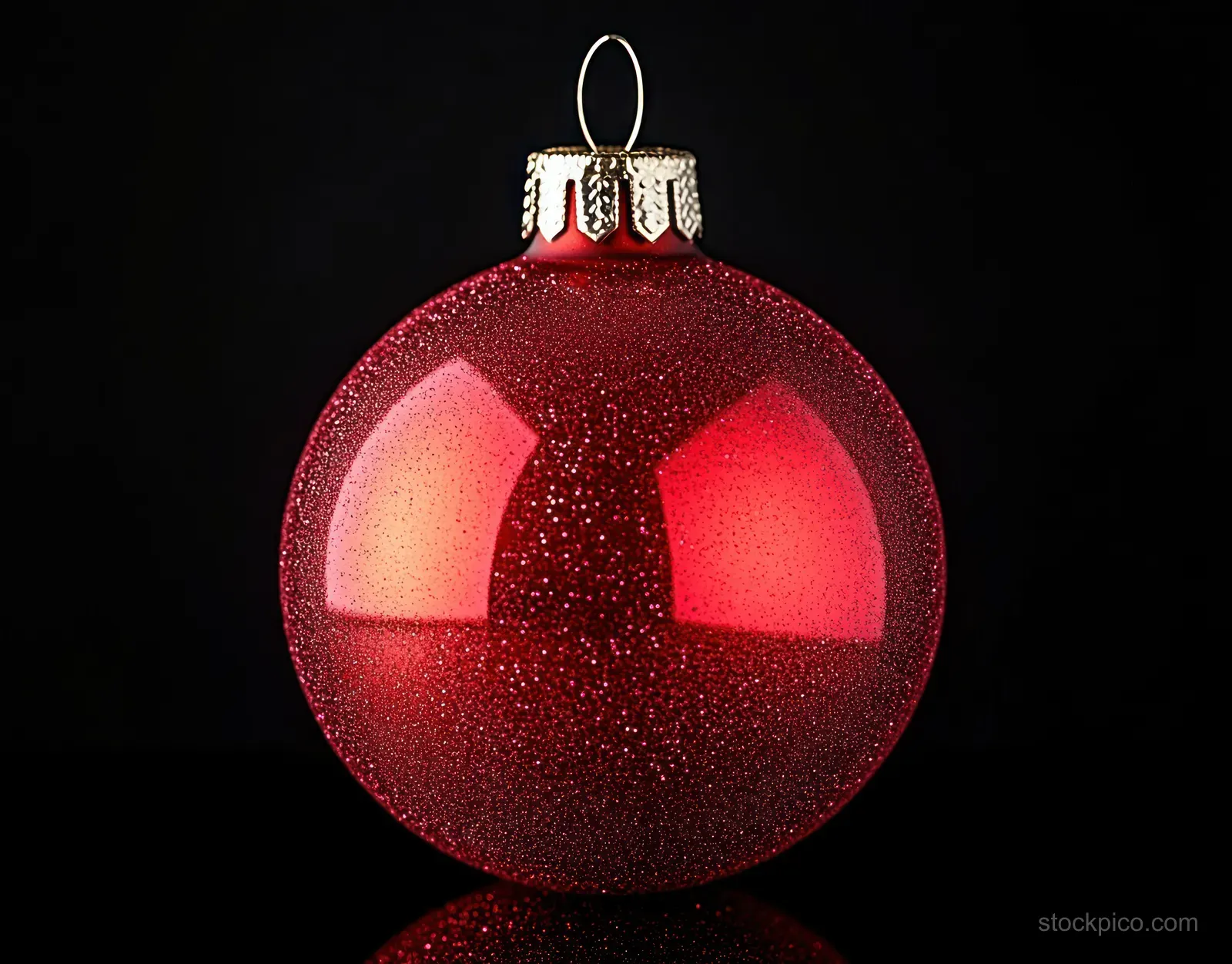 A red glitter Christmas ornament ball with a golden cap and hanging hook reflected on a glossy black background.
