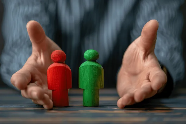 Hands split two wooden figures, one red and one green, on a table to symbolize mediation.