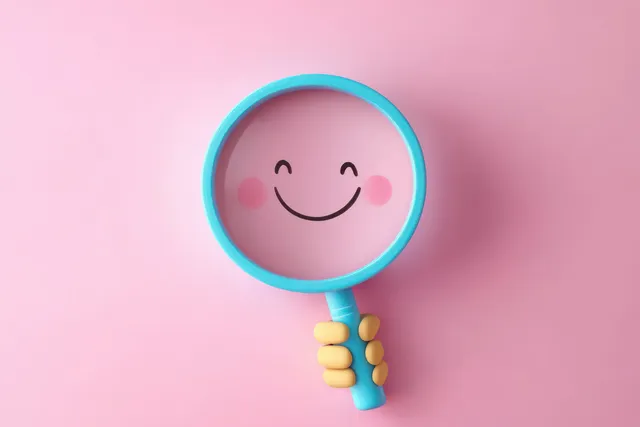 Yellow hand holds a blue magnifying glass revealing a smiling face on pink background
