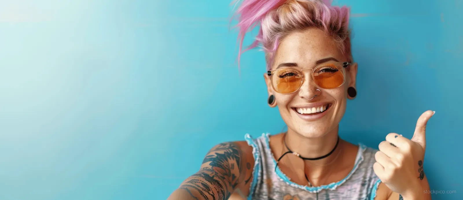 Smiling pink-haired woman with tattoos gives a thumbs-up while wearing sunglasses against a blue backdrop.