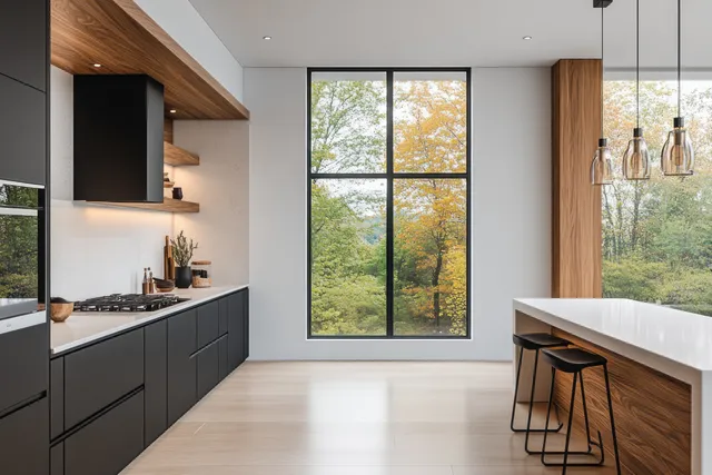 Minimalist kitchen interior with black cabinets, white counter, warm wood shelves, and a large window showing autumn trees.