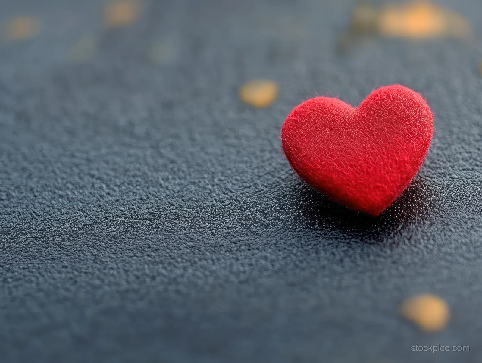 Small red heart-shaped ornament resting on a dark textured surface with soft blurred lights in the background.