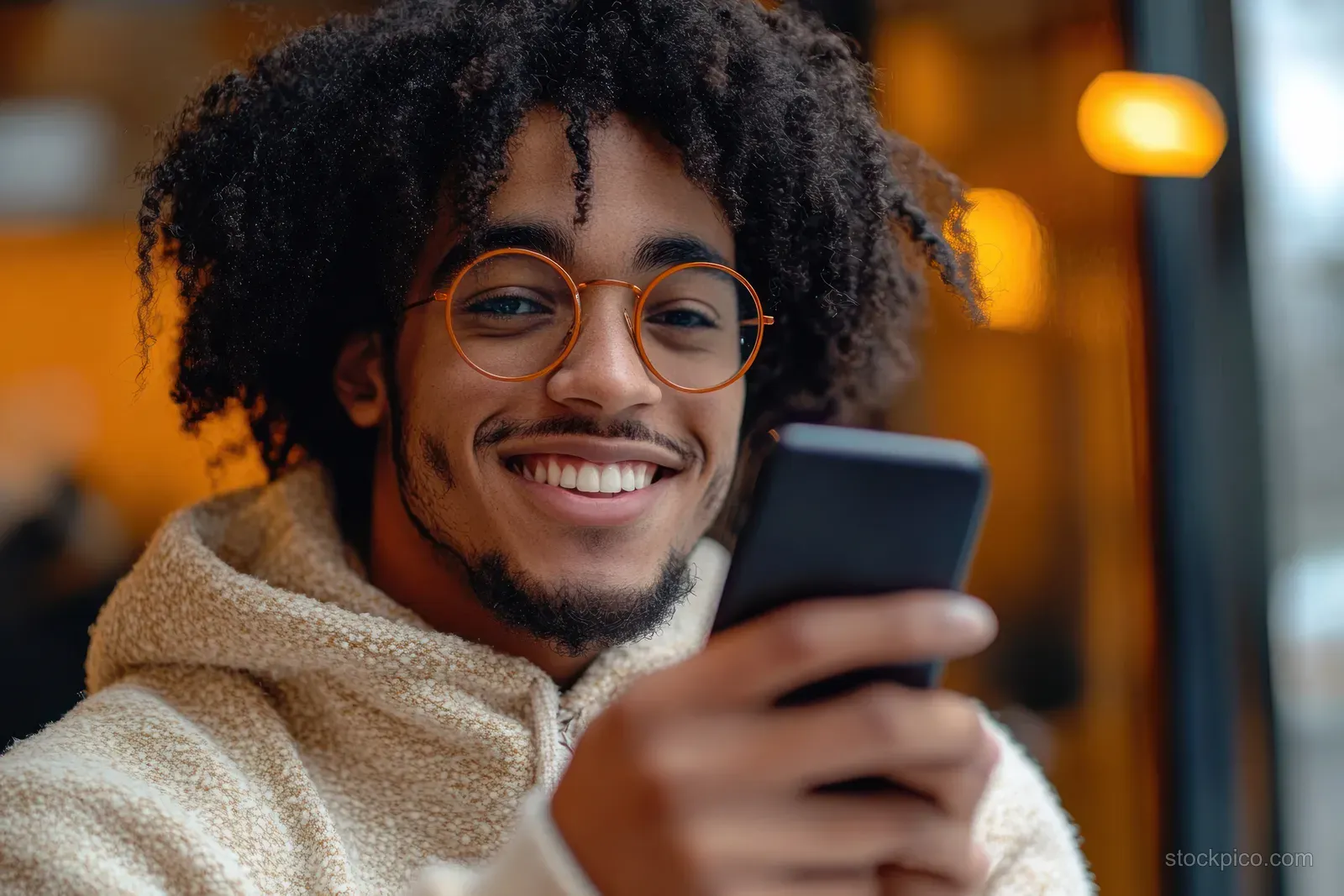 A smiling young man with curly hair and round glasses holds a smartphone indoors in warm ambient light.