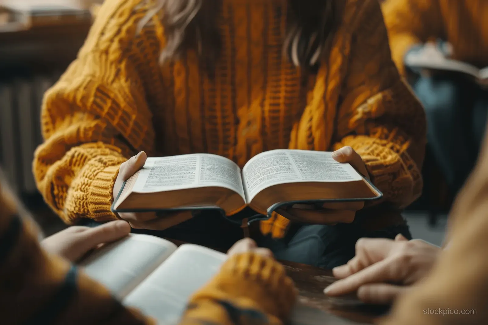 Hands hold an open Bible while a Christian group reads together in a sunlit study room.