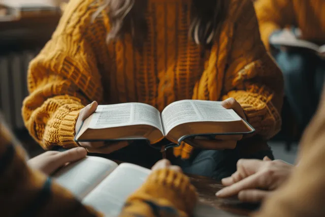 Hands hold an open Bible while a Christian group reads together in a sunlit study room.