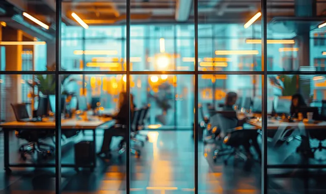 Blurred office floor through glass panels with warm lights and working desks at sunset