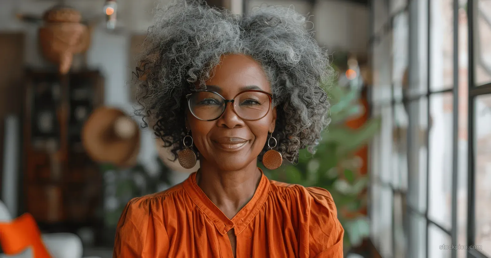 A smiling senior woman with curly hair and glasses holds a smartphone while standing indoors.