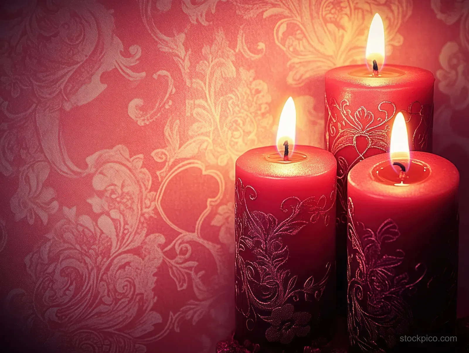 Three red candles with floral designs glow against patterned red wallpaper with warm flickering light.