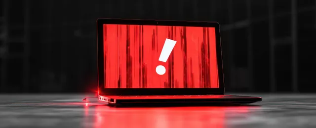 A black laptop shows a bright red error screen with a large white exclamation mark on a dark surface.