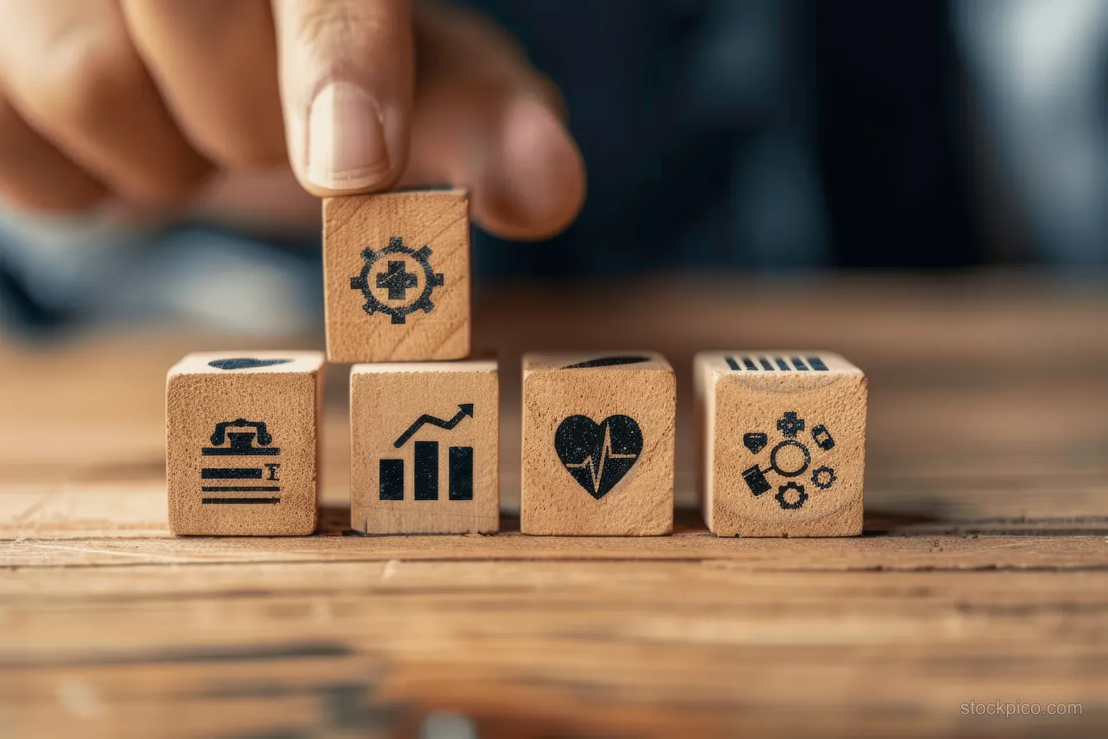 A hand places wooden blocks with icons for health insurance, pensions, and benefits on a desk.
