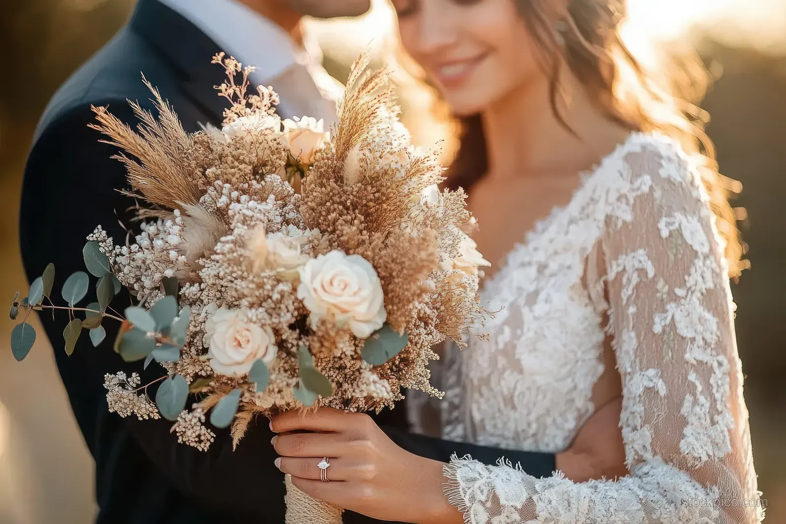 Bride and groom embrace outdoors as the bride holds a rustic dried flower bouquet with white roses in golden light.