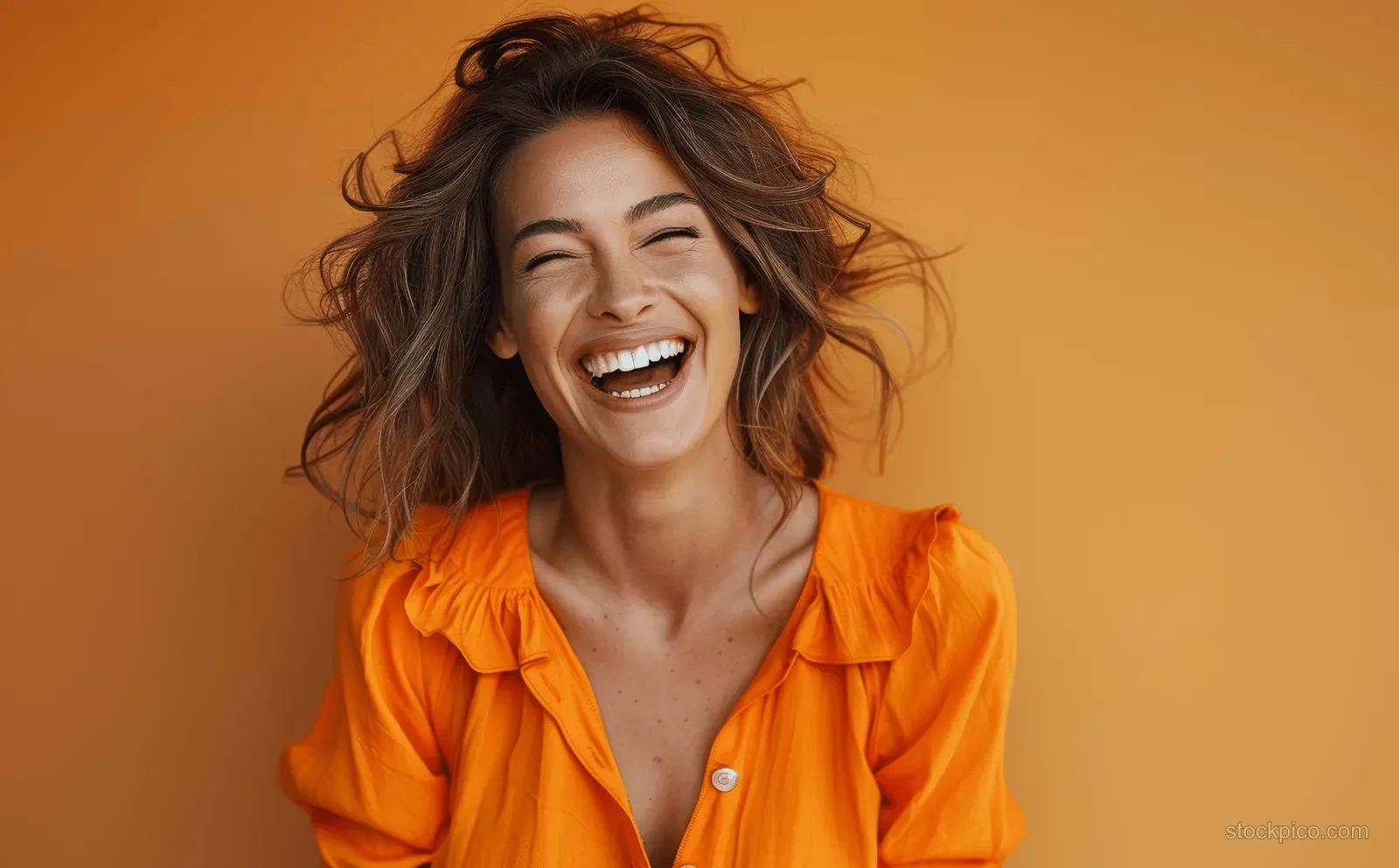 A cheerful businesswoman laughs in a studio portrait wearing a bright orange blouse against a warm orange background.