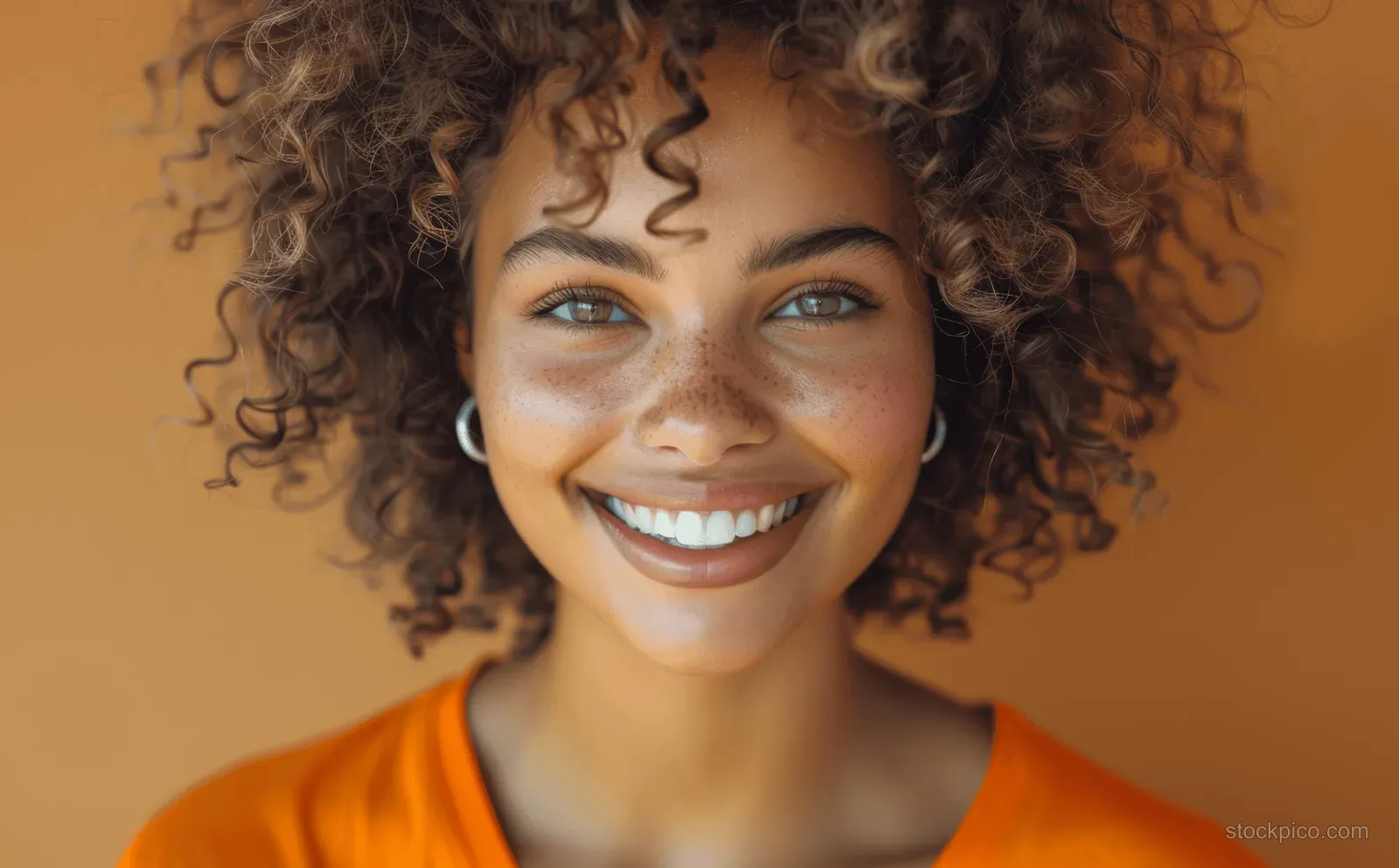Smiling young woman with curly hair wearing an orange shirt against a brown background.