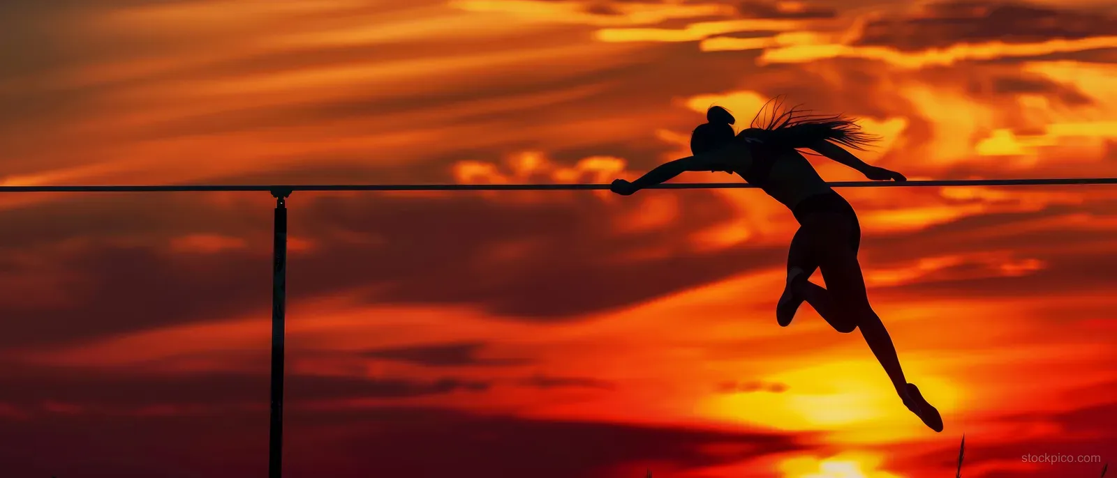 A silhouetted high jumper clears the bar on a stadium track against a vivid sunset sky.