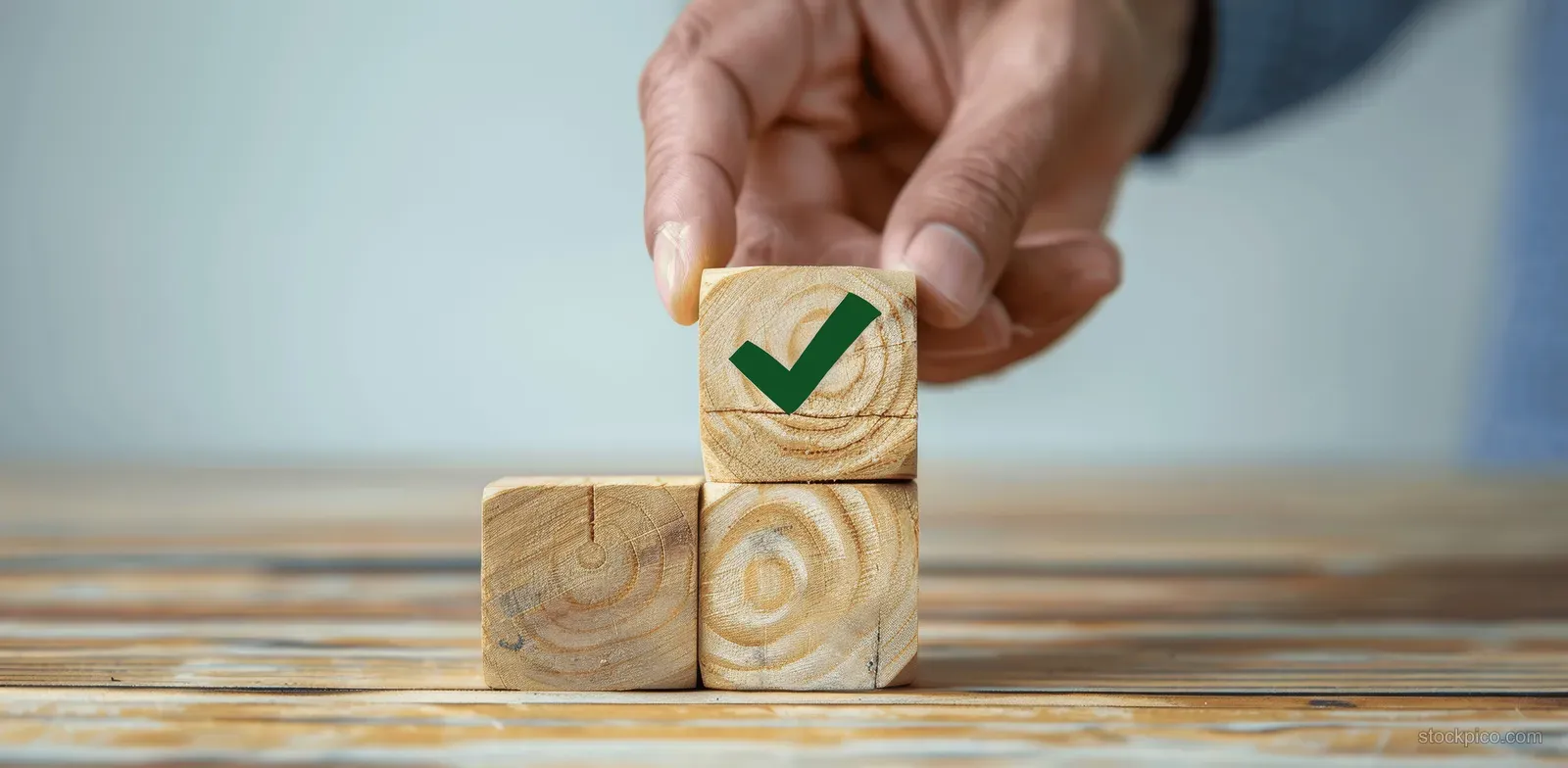 A hand presses a green check mark onto stacked wooden blocks indicating quality compliance.