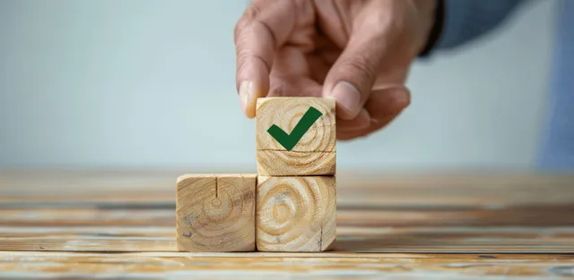 A hand presses a green check mark onto stacked wooden blocks indicating quality compliance.