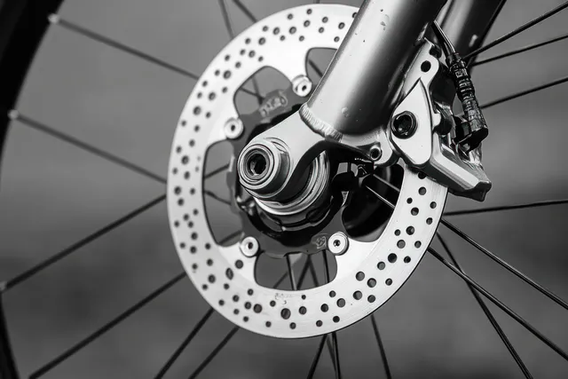 Bicycle front wheel disc brake rotor and caliper are shown in close-up with spokes fading into a soft background.
