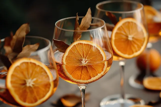 Close-up of wine glasses filled with amber liquid, garnished with dried orange slices and delicate leaves in warm autumn tones.