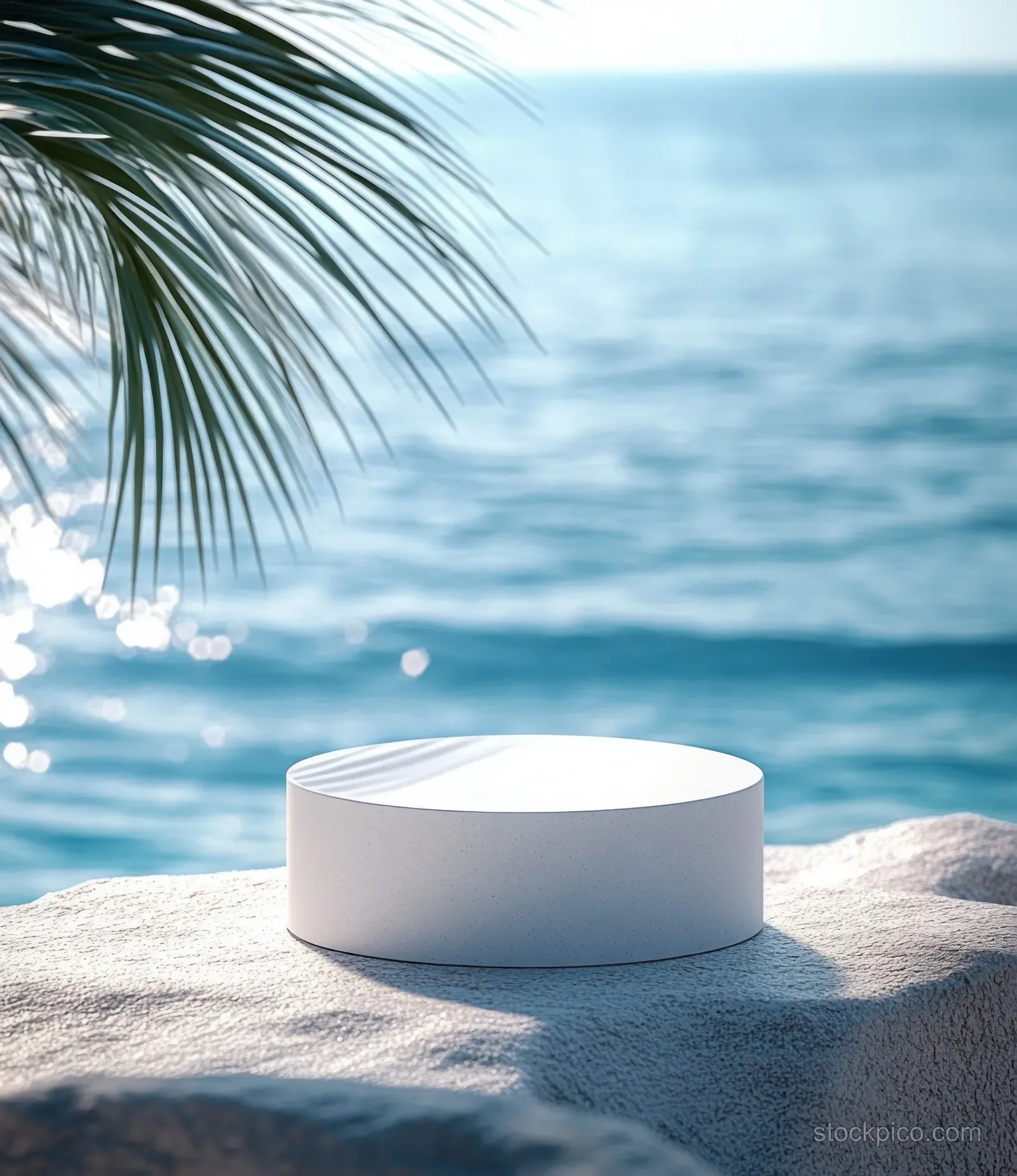 A white round pedestal sits on a textured stone surface beside palm leaves and a calm ocean.