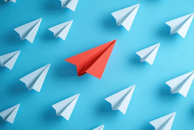 Red paper airplane leads among white planes on blue background, aiming forward