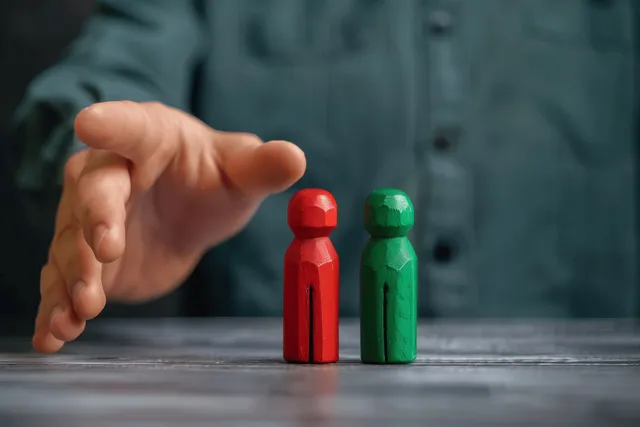 A hand gestures beside two standing red and green wooden figures on a table.
