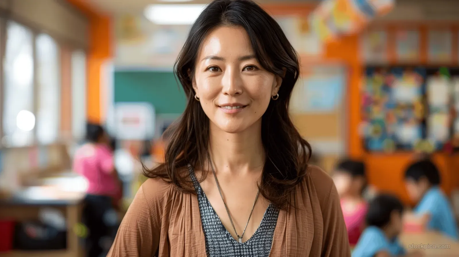 Asian female teacher smiles with hands on hips in a colorful preschool classroom as children play behind her.