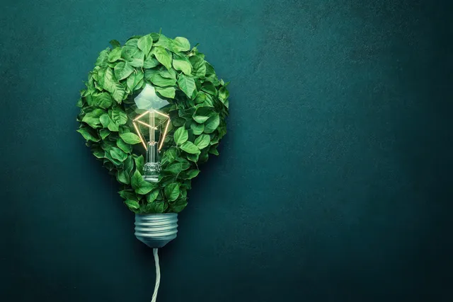 A green leaf-shaped light bulb with a glowing filament on a dark textured background representing eco energy.