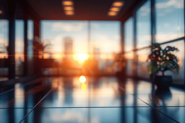 Blurred office interior with large panoramic windows and warm sun flare over desks and chairs.