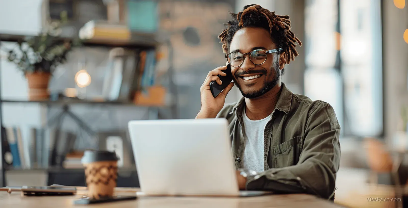 Smiling young man talking on a smartphone beside an open laptop at an office desk.