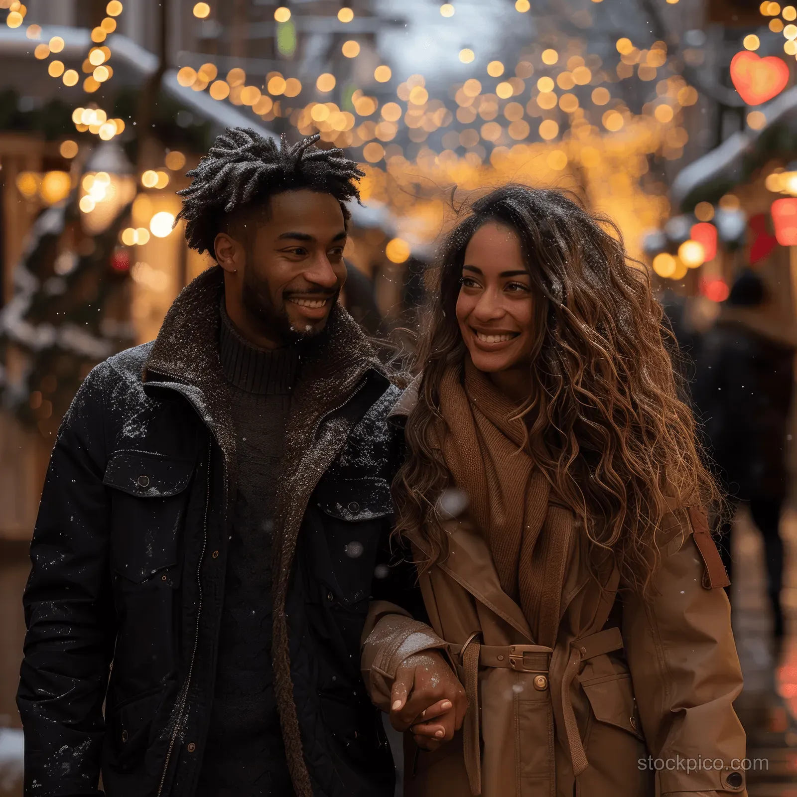 A smiling couple walk hand in hand on a snowy Christmas street with glowing holiday lights behind them.