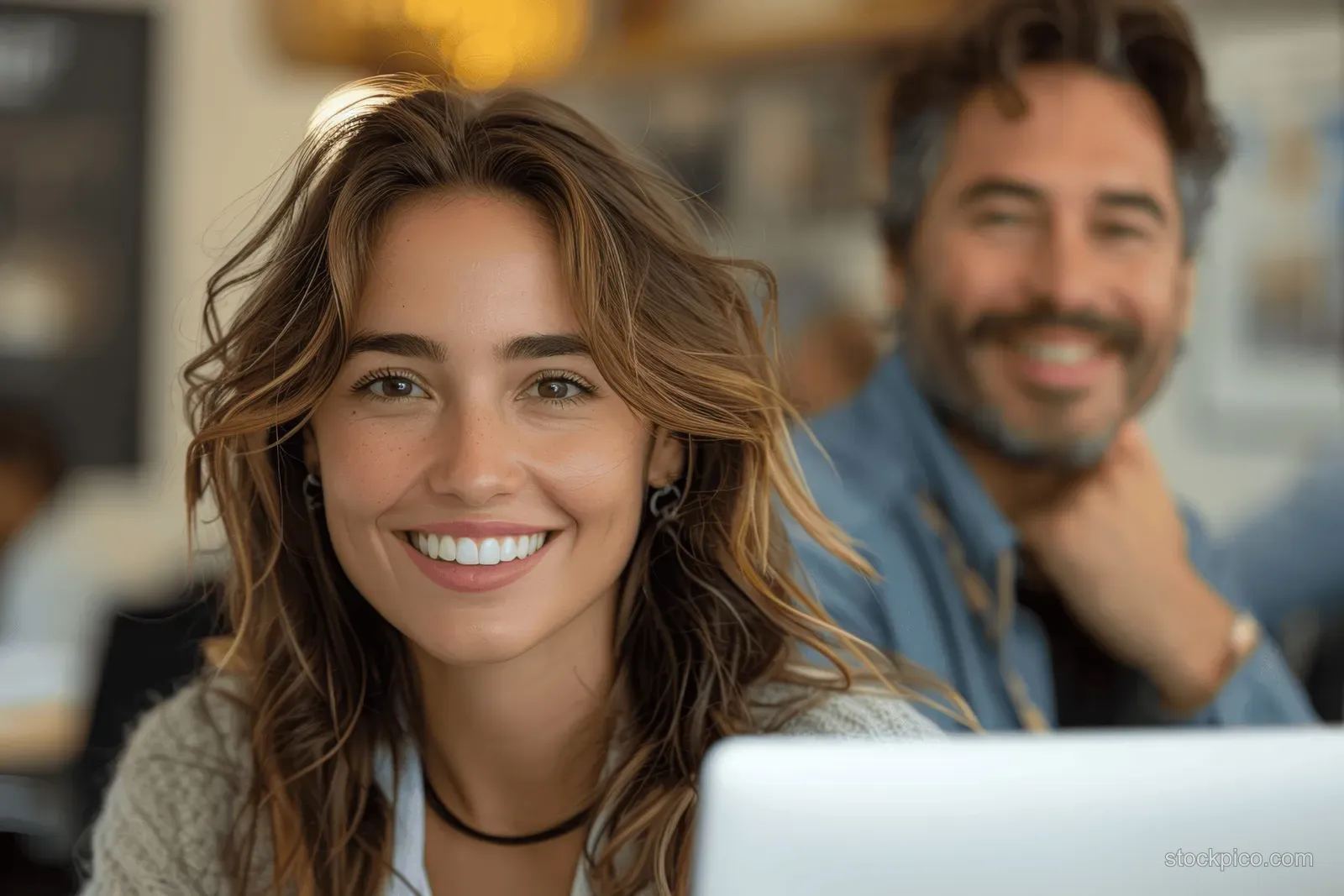 Smiling businesswoman in foreground with a male colleague using a laptop in a bright office.