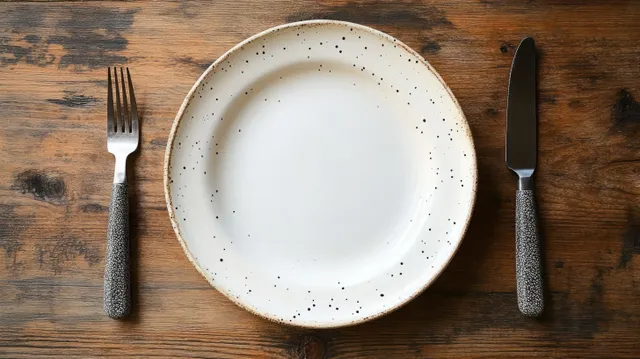 Empty white speckled ceramic plate on a rustic wooden table with a fork and knife beside it.