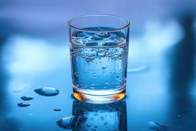 Clear glass of sparkling water with ice cubes on a reflective blue surface, surrounded by condensation and water droplets.