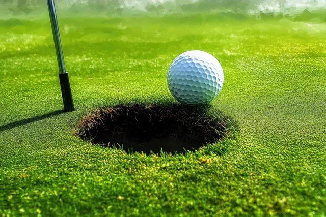Golf ball rests on the rim of a hole on a bright putting green with a putter nearby casting a shadow.