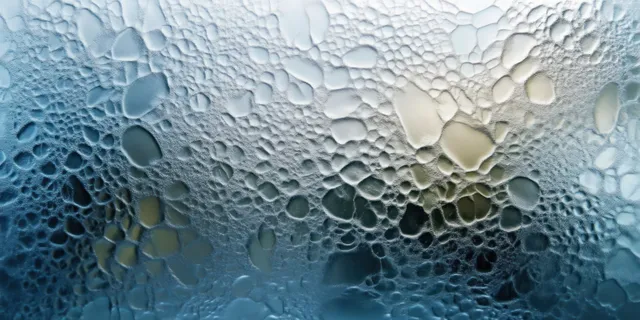 Water droplets cover a glass pane in a textured condensation pattern with a blurred cool background.