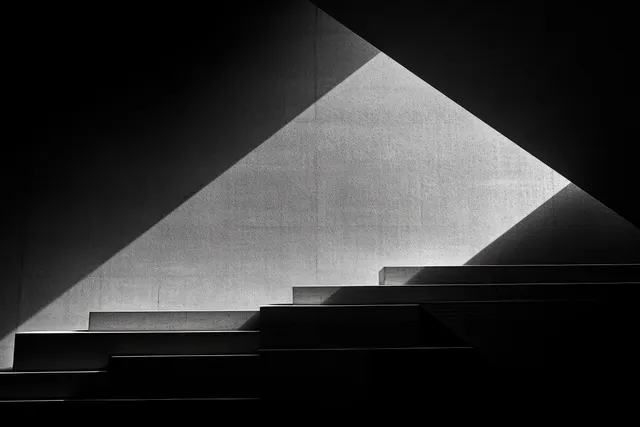 Triangular shadows and light patterns stretch across a stepped concrete interior in black and white.