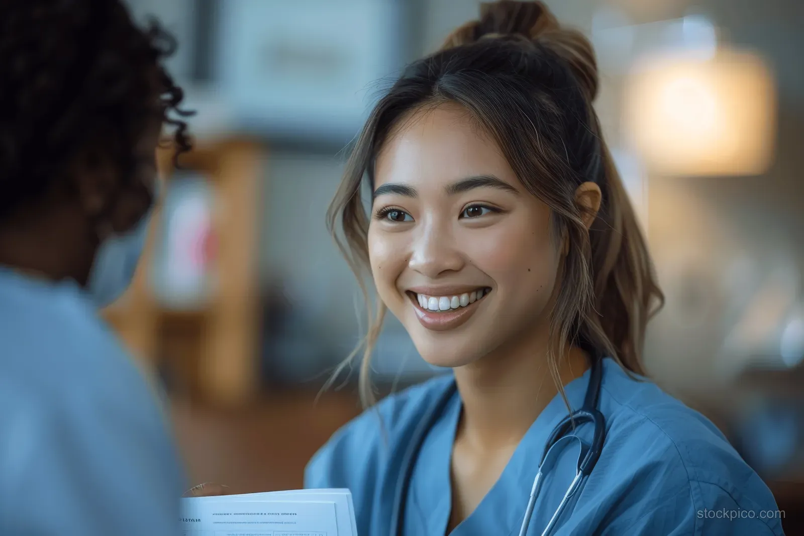An Asian nurse smiles while greeting a black female patient in a softly lit hospital hallway.