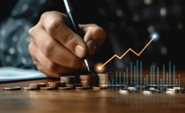 A hand draws an upward stock chart on a desk beside scattered coins and a glowing market dashboard.