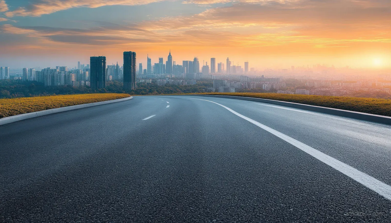 Curved asphalt road leading toward a distant city skyline at sunset.
