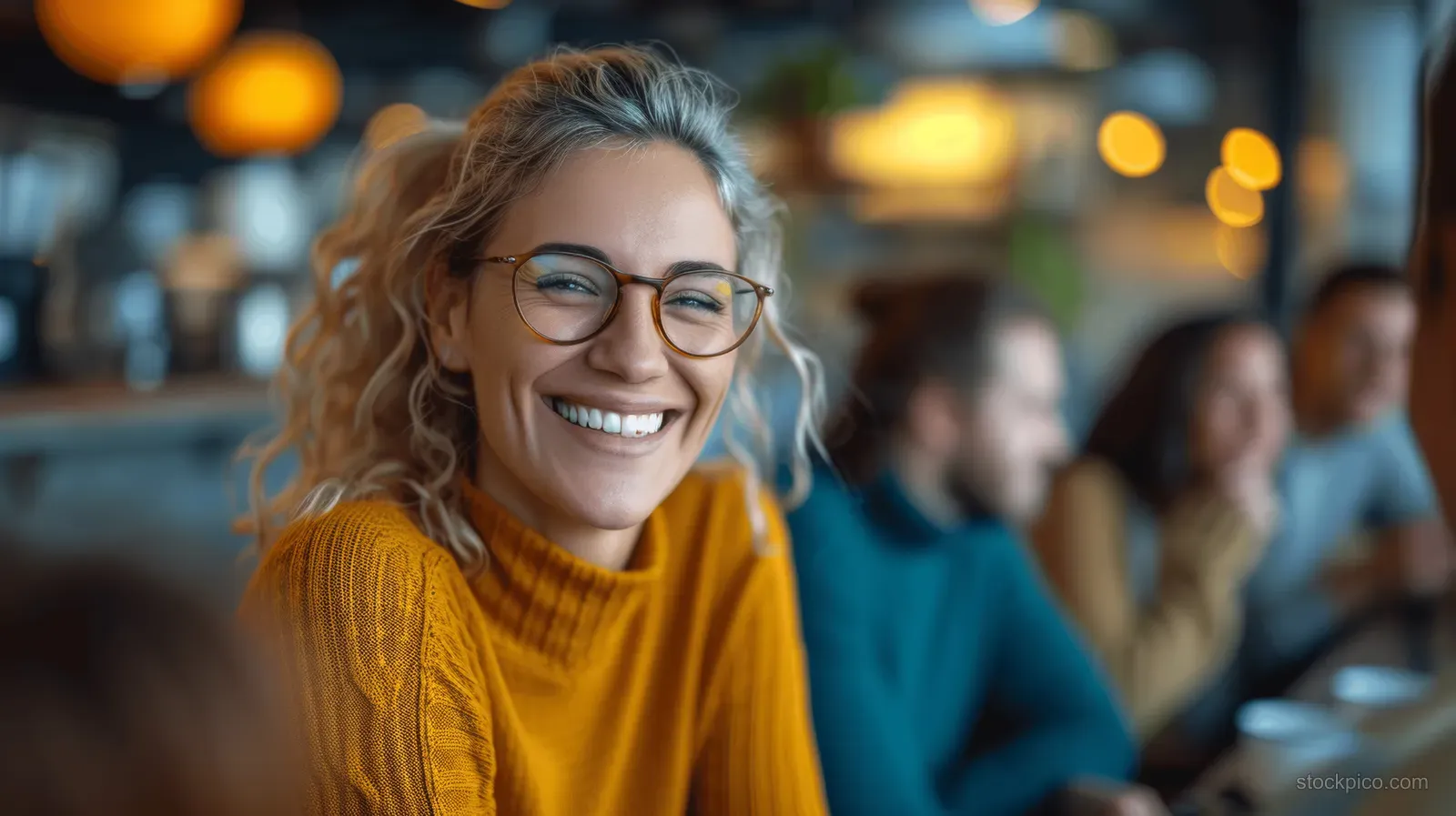 Smiling woman with glasses in a yellow sweater sits in a café with coworkers blurred behind her.