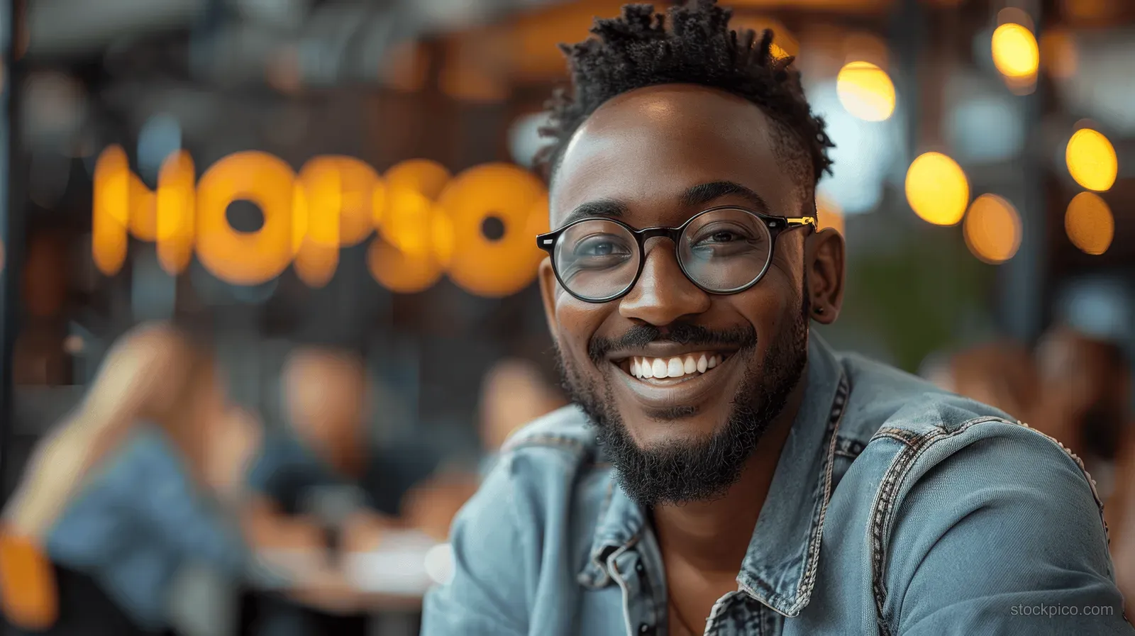 Smiling man wearing glasses and a denim jacket seated in a warmly lit cafe with blurred bokeh lights behind.