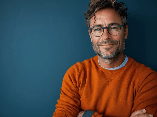 A smiling man in an orange sweater and glasses poses with arms crossed against a solid blue background.