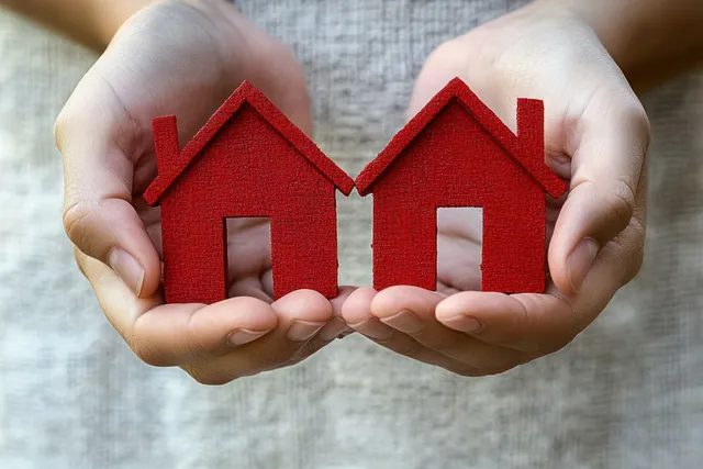 Two hands hold two separated red house symbols on fabric, suggesting divorce and property division.