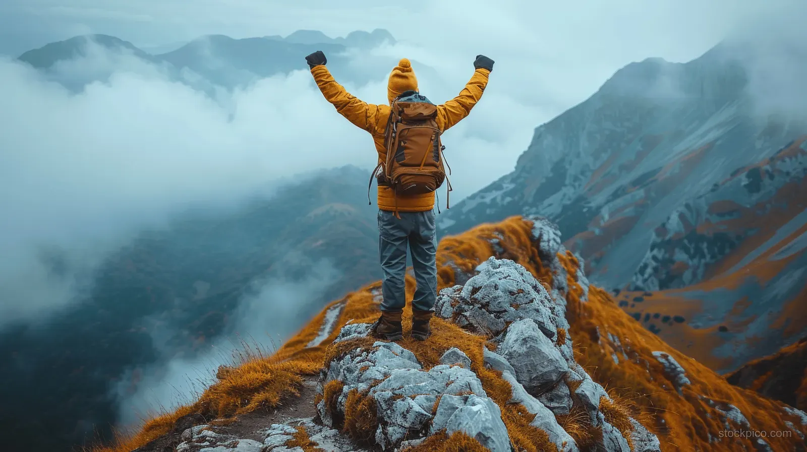 A hiker celebrates with arms raised on a rocky mountain peak above a fog-filled valley at sunrise.
