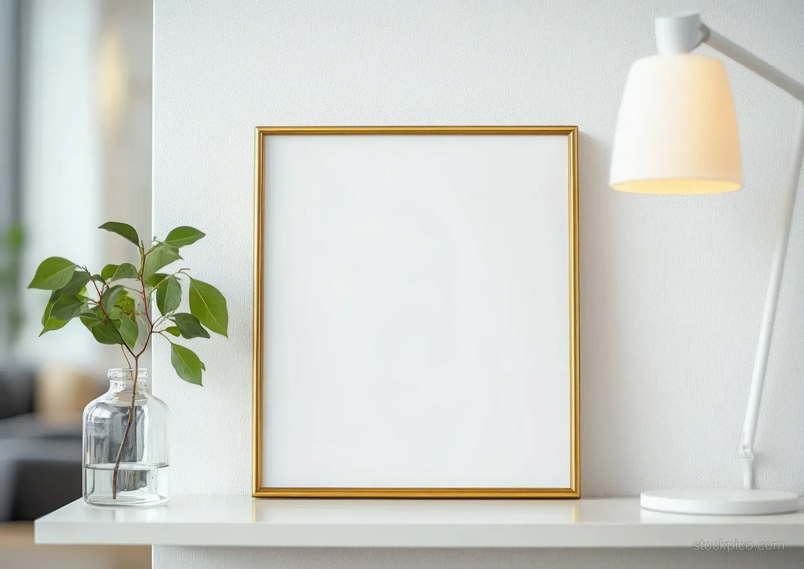 Gold picture frame centered on a white shelf beside a glass vase with green twig and a modern desk lamp.