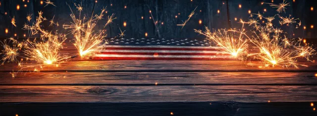 Sparklers burn on a rustic wooden table in front of American flag stripes with warm spark bokeh.