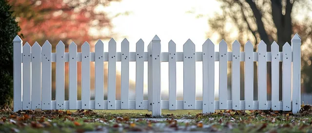 White pointed picket fence in a leaf-covered yard with blurred trees and bright autumn sunlight behind.
